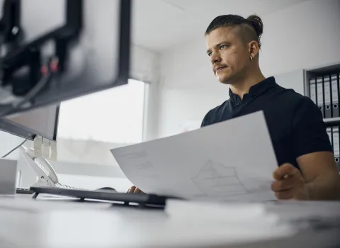 Ein junger technischer Systemplaner sitzt an einem Schreibtisch und hält einen Bauplan in den Händen.