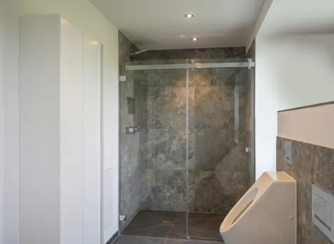 Eleganter Duschbereich mit Natursteinoptik, integrierter Ablage, Chromarmatur und angrenzendem Urinal.