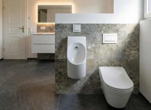 Eleganter Sanitärbereich mit Urinal, WC, Natursteinwand, beleuchtetem Spiegel und goldfarbenem Türgriff.