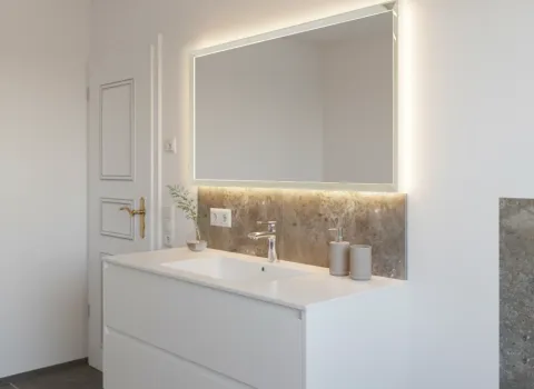 Hochwertiger Waschplatz mit weißem Möbelstück, Lichtspiegel, goldfarbenem Türgriff und klarer Linienführung.