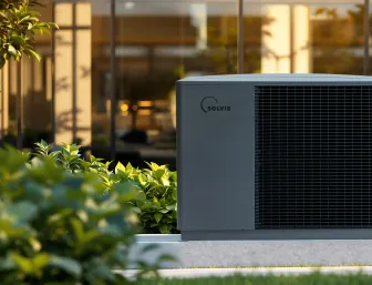 Eine Solvis-Wärmepumpe steht im begrünten Außenbereich vor einem modernen Gebäude, umgeben von Pflanzen und warmem Sonnenlicht.