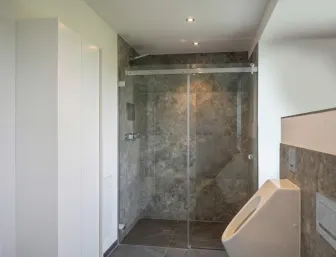 Eleganter Duschbereich mit Natursteinoptik, integrierter Ablage, Chromarmatur und angrenzendem Urinal.