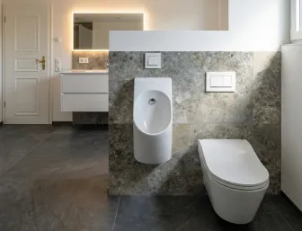 Eleganter Sanitärbereich mit Urinal, WC, Natursteinwand, beleuchtetem Spiegel und goldfarbenem Türgriff.